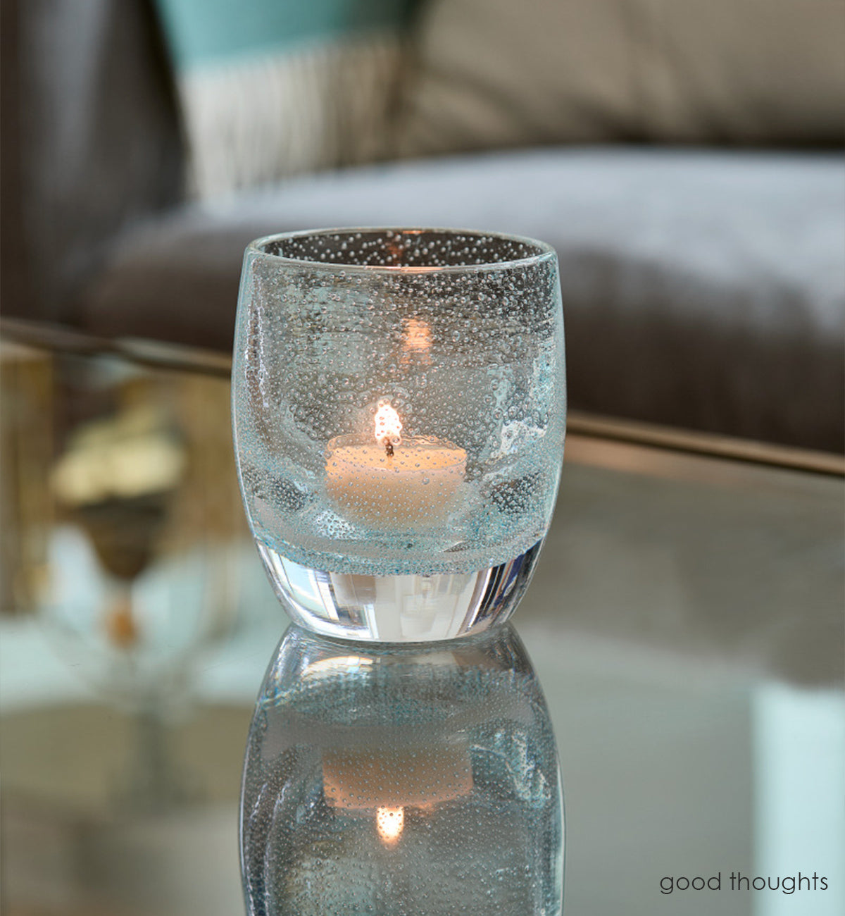 good thoughts, transparent light blue with small bubble pattern hand-blown glass votive candle holder on a reflective coffee table with gray couch in background.