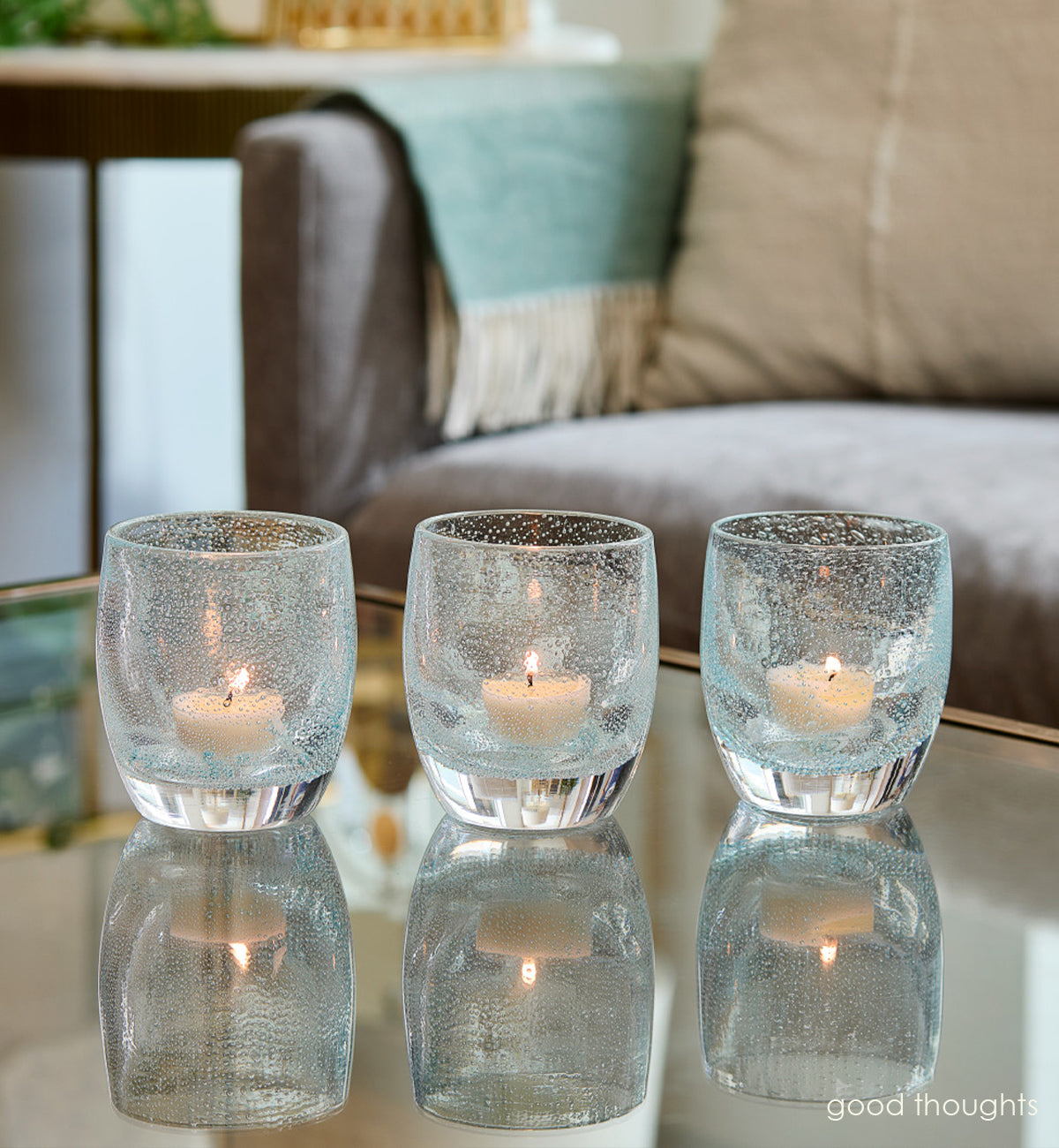 three good thoughts, transparent light blue with small bubble pattern hand-blown glass votive candle holders on a reflection coffee table with couch and side table in background.