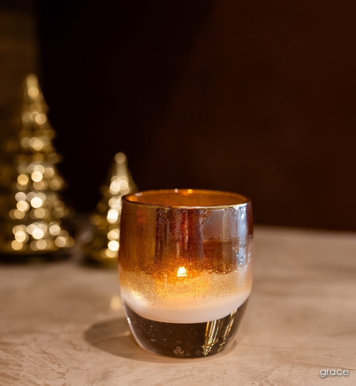 grace golden topped white hand-blown glass votive candle holder with gold trees behind it.