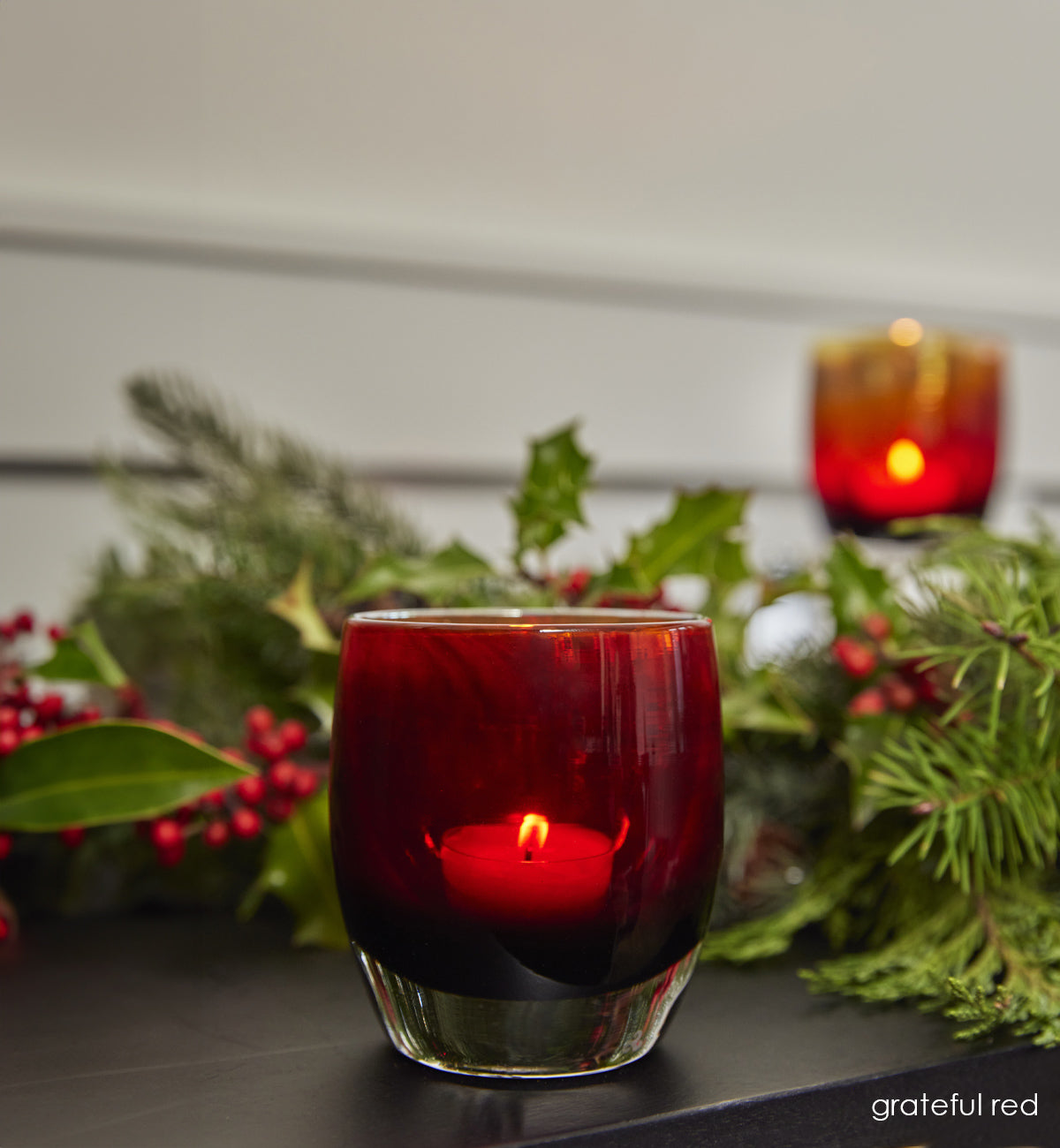 two grateful red wine red with metallic interior hand-blown glass votive candle holders with lit tealights on a dark table with holly berries.