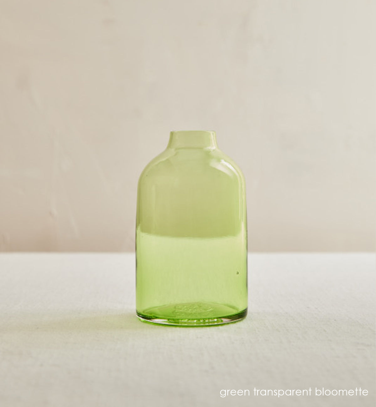 green transparent bloomette, transparent green hand-blown glass flower bud vase on a cream table in front of cream wall.