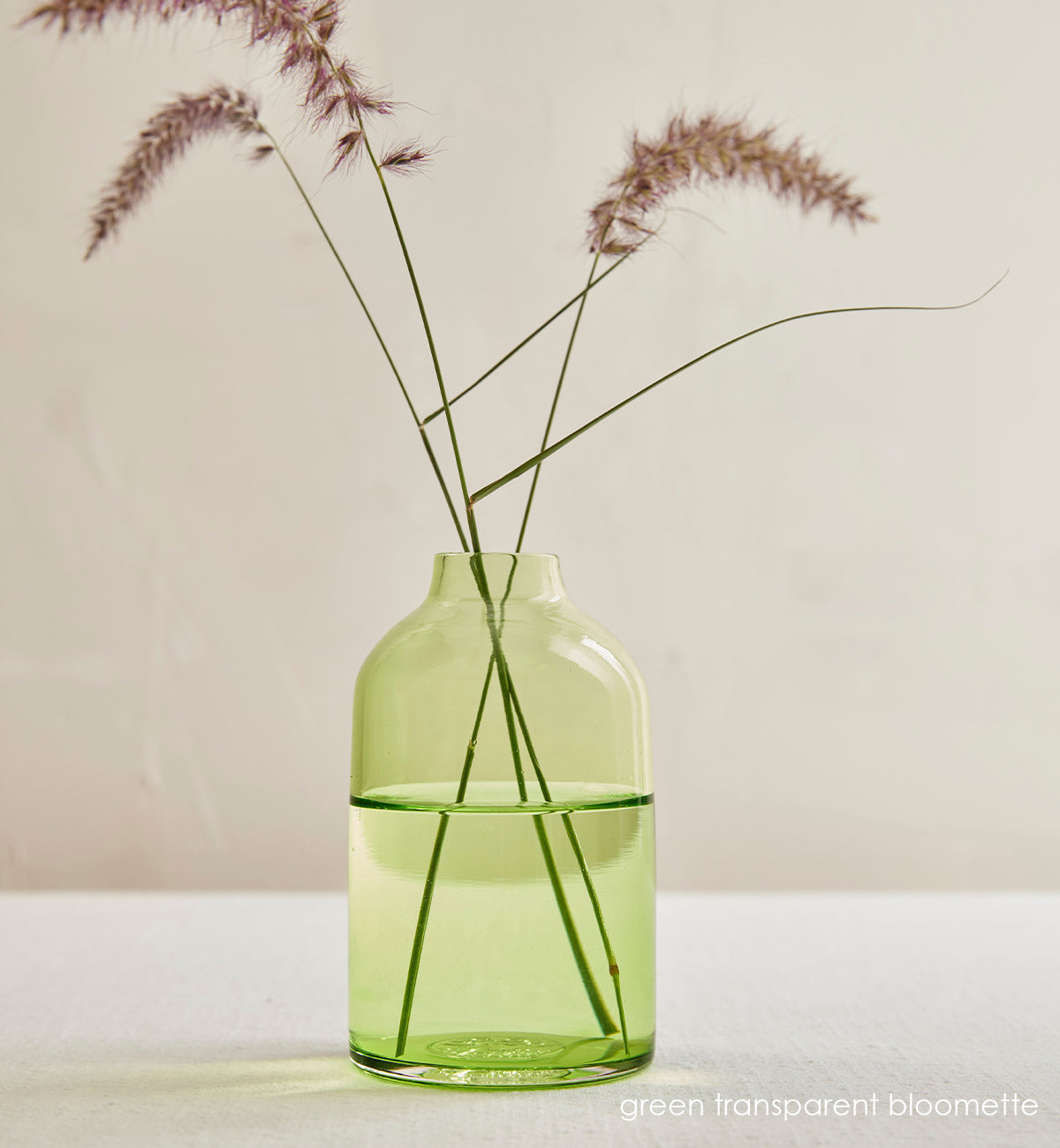 green transparent bloomette, transparent green hand-blown glass flower bud vase with purple grass inside in an interior cream setting.