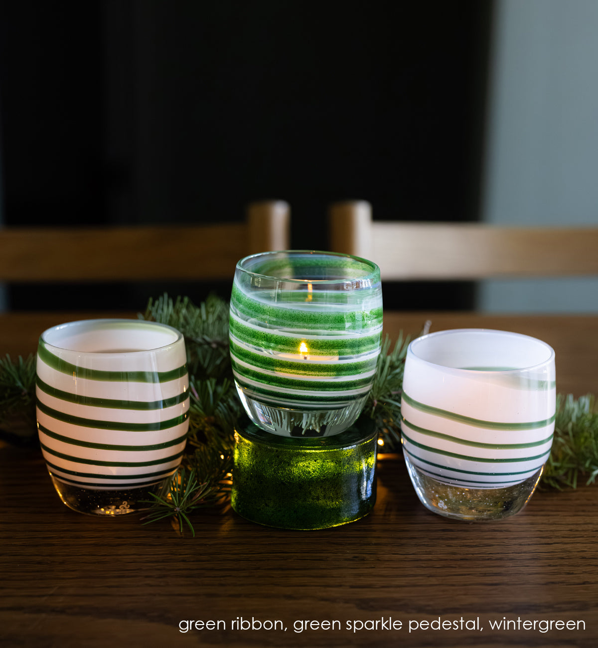 'green ribbon' glassybaby: green, white, clear swirl handblown glass candle holder sitting on green sparkle pedestal with two wintergreen glassybaby on the sides sitting on wood table with garland