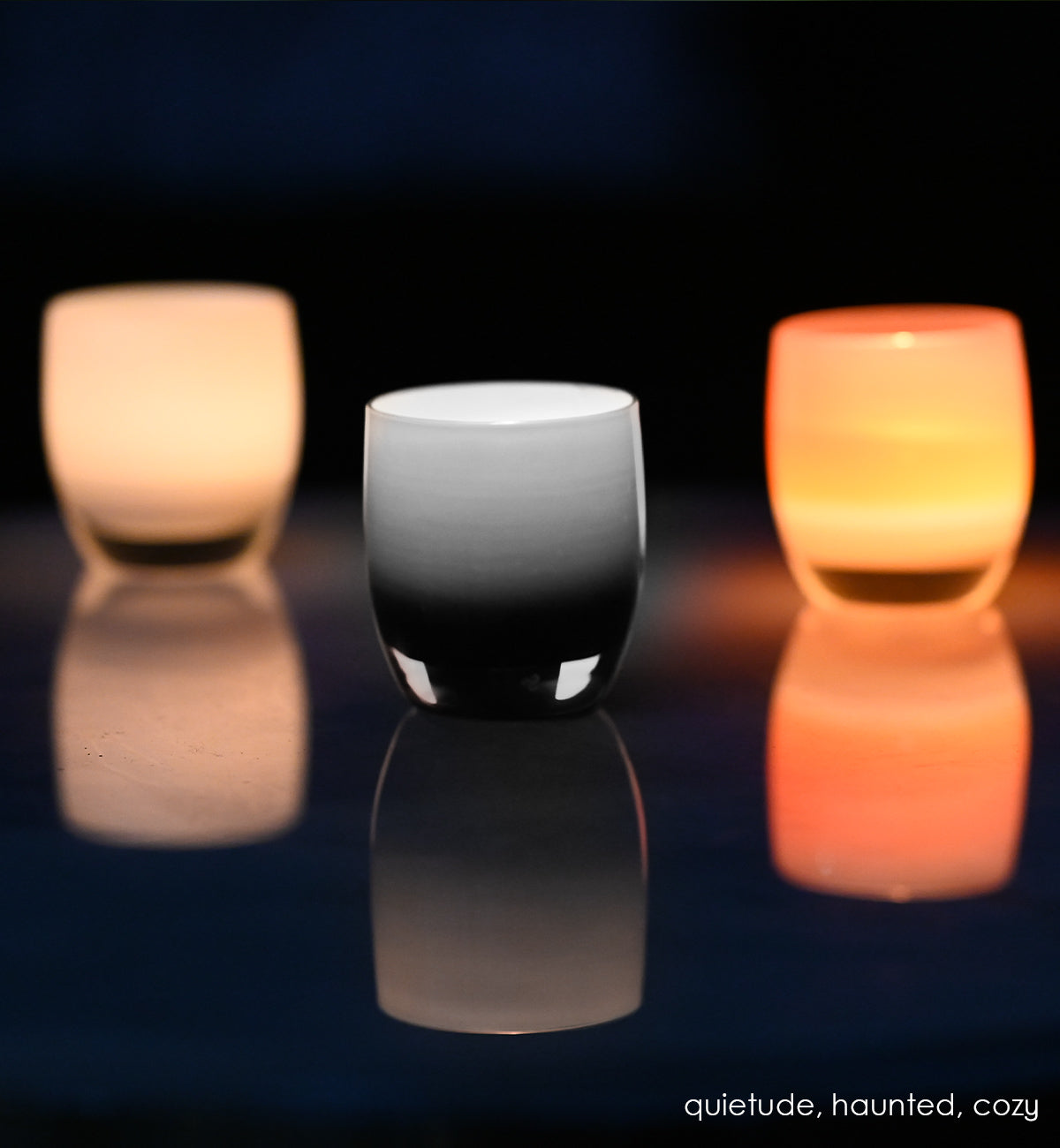 haunted black double overlay hand-blown glass votive candle holder with quietude and cozy all lit with tealights on a reflective surface.