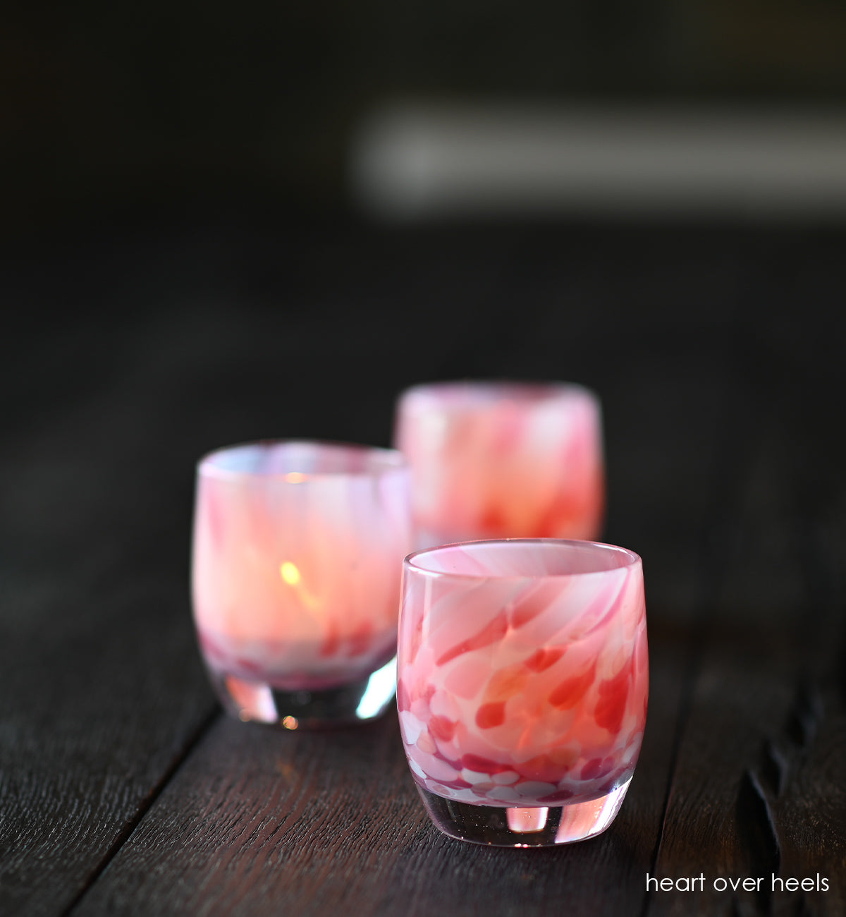 three heart over heels white and pink speckled hand-blown glass votive candle holders on a charcoal wood table.