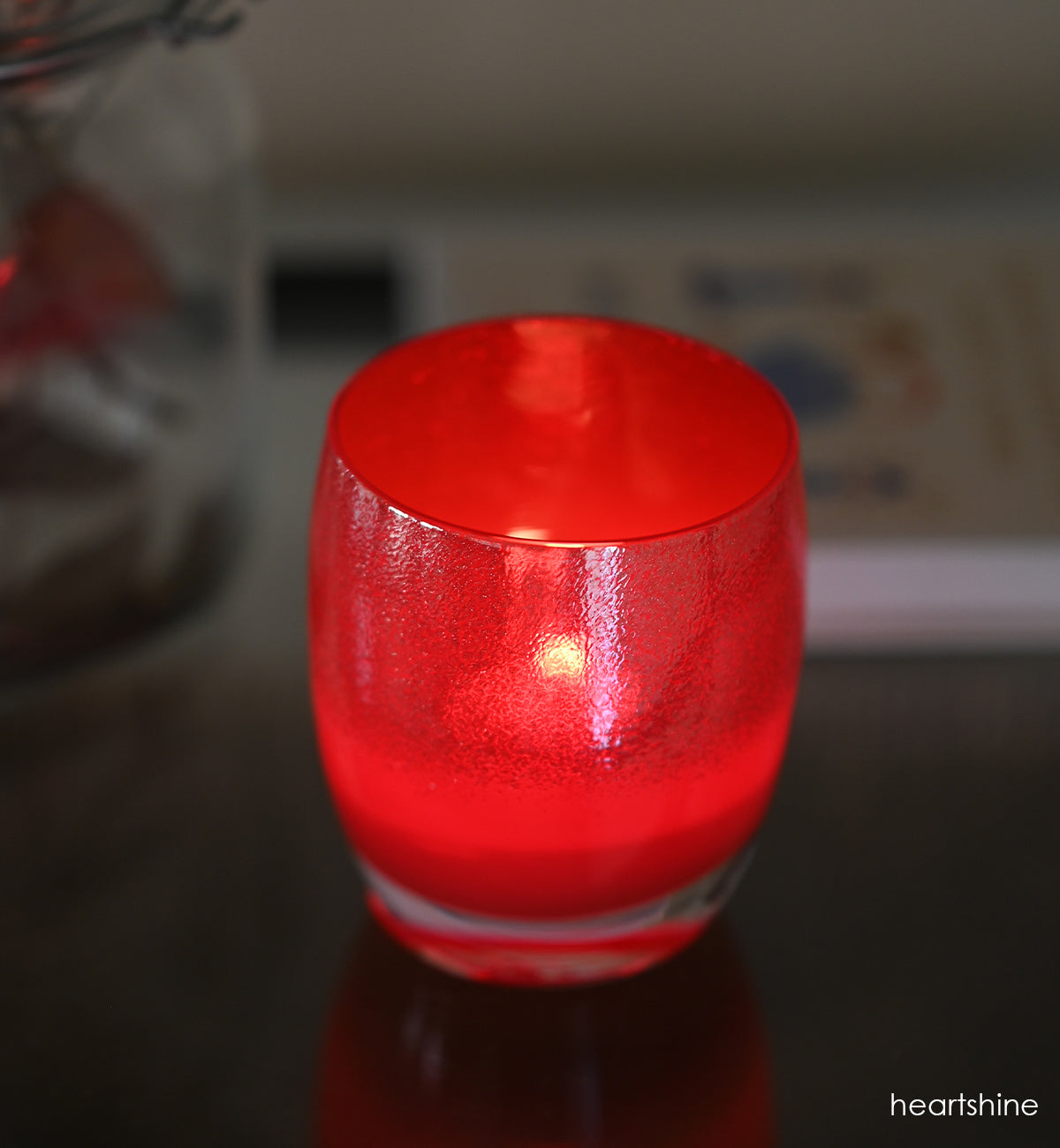 heartshine, shimmering glitter that enrobes the top of a bright red base, hand-blown glass votive candle holder lit in a dim setting.
