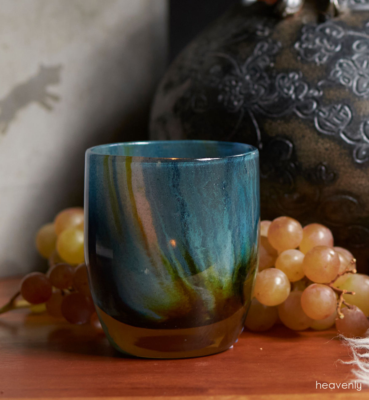 heavenly, blue green swirl, hand-blown glass votive candle holder atop a table with a vase and grapes.