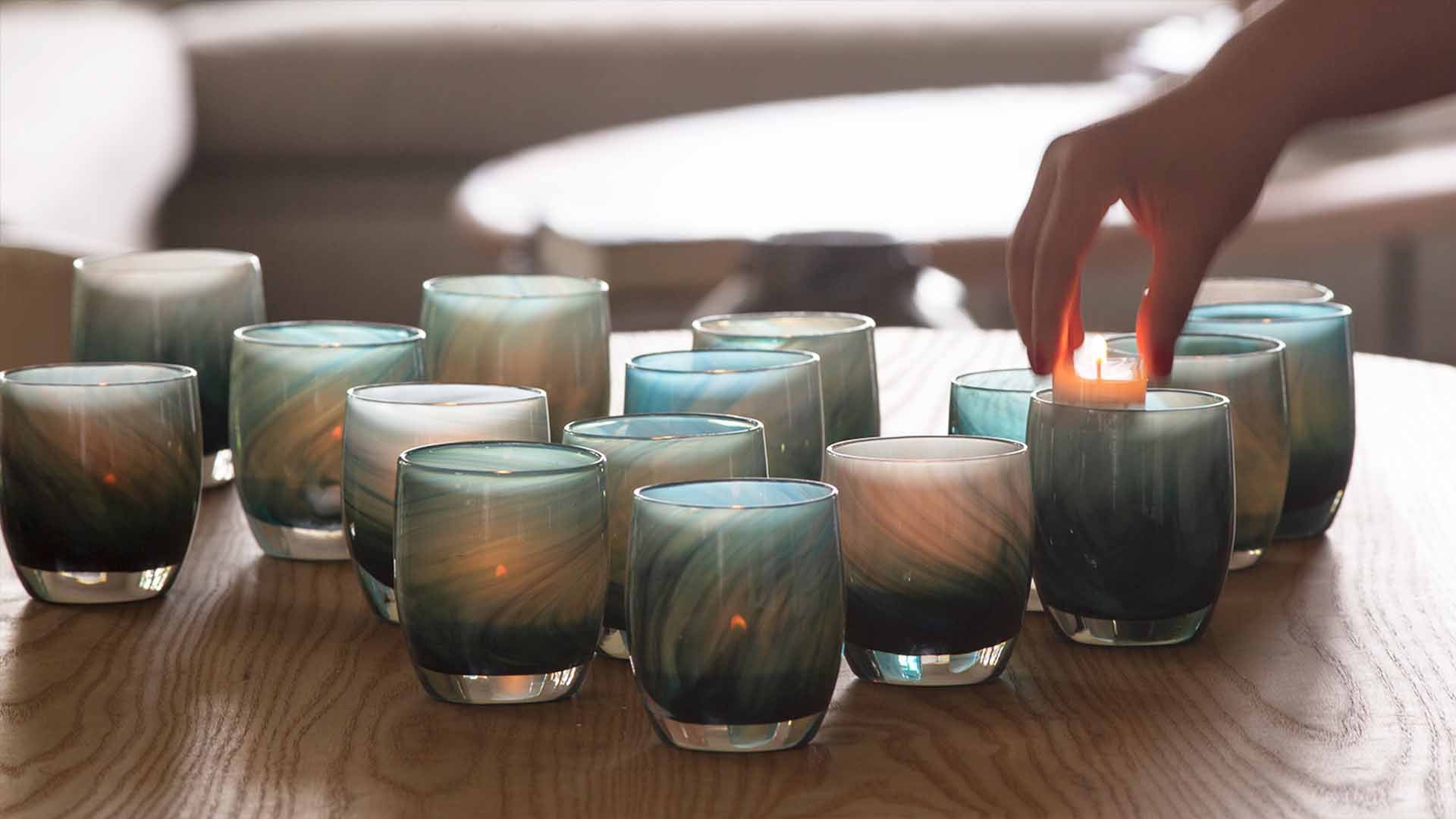 Hand dropping a lit tealight into a collection of homerun glassybaby votives on a wood table. 