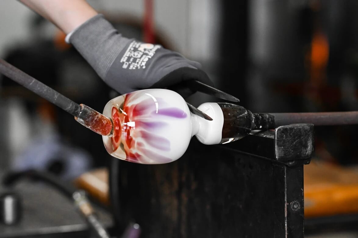glassblower making petal, white with pink and purple petals hand-blown glass votive candle holder in a hot shop.