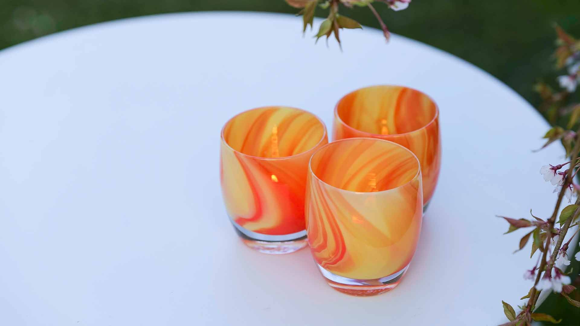 Three lifesaver sitting grouped on a round white table in the garden. Marbled orange hand-blown glass votive candle holder.