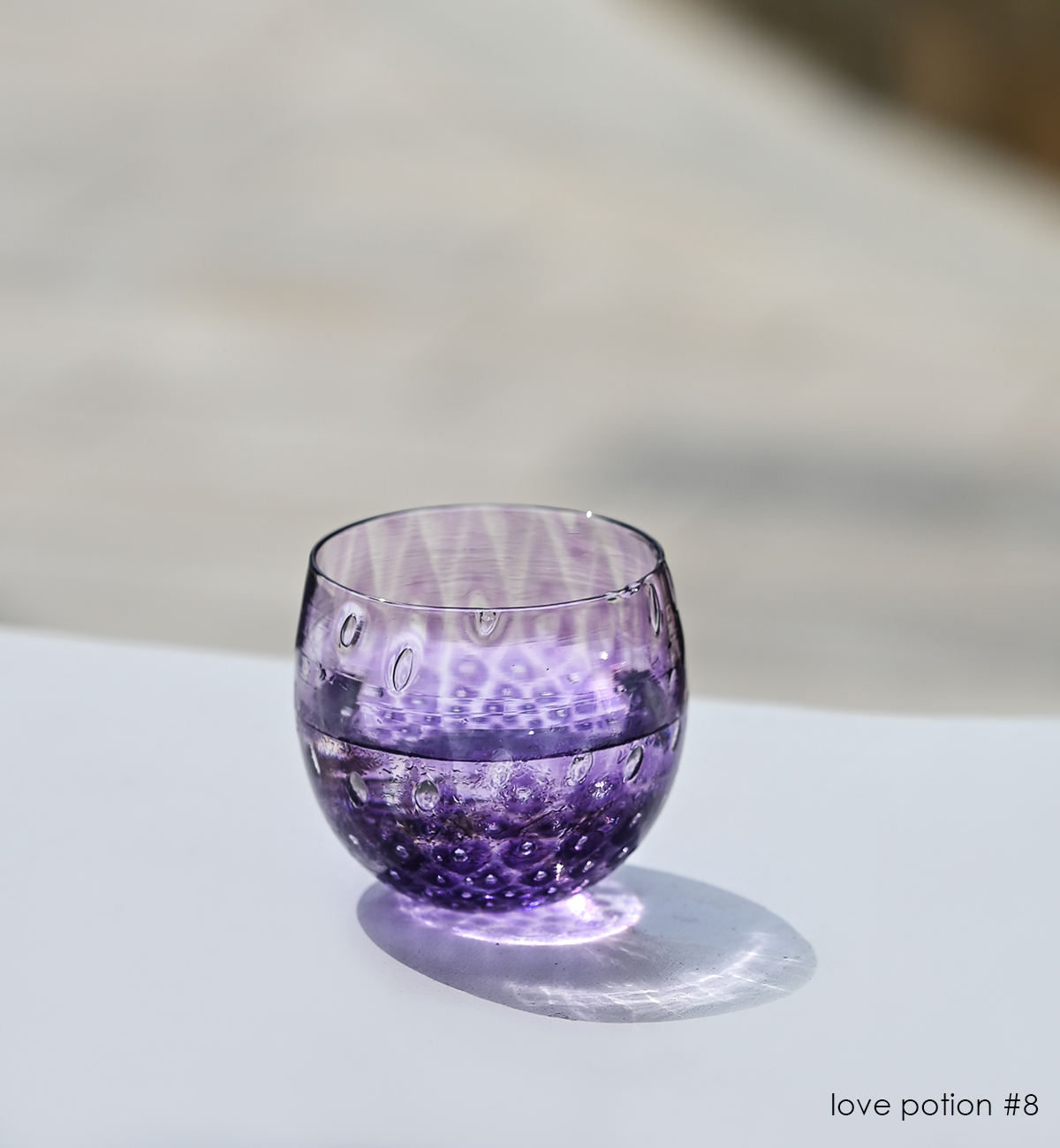 love potion #8, a deep purple hand-blown drinking glass with a bubble pattern, sitting on white surface in sunlight.