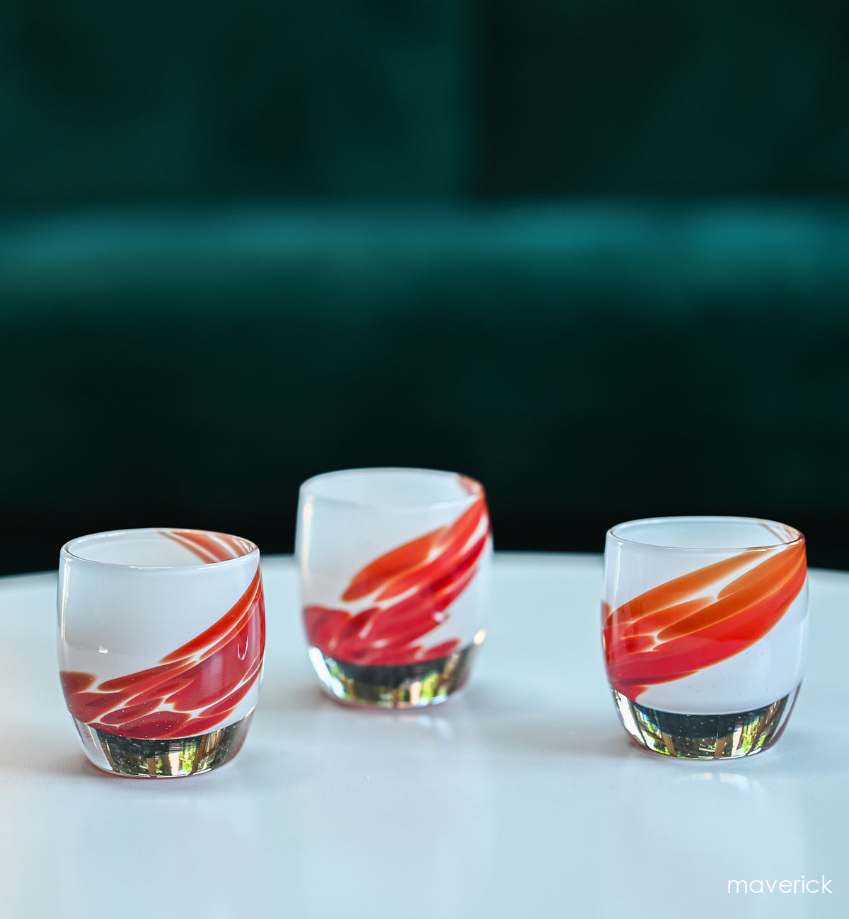 three maverick white with red wing, hand-blown glass votive candle holders. on a white table with green couch behind.