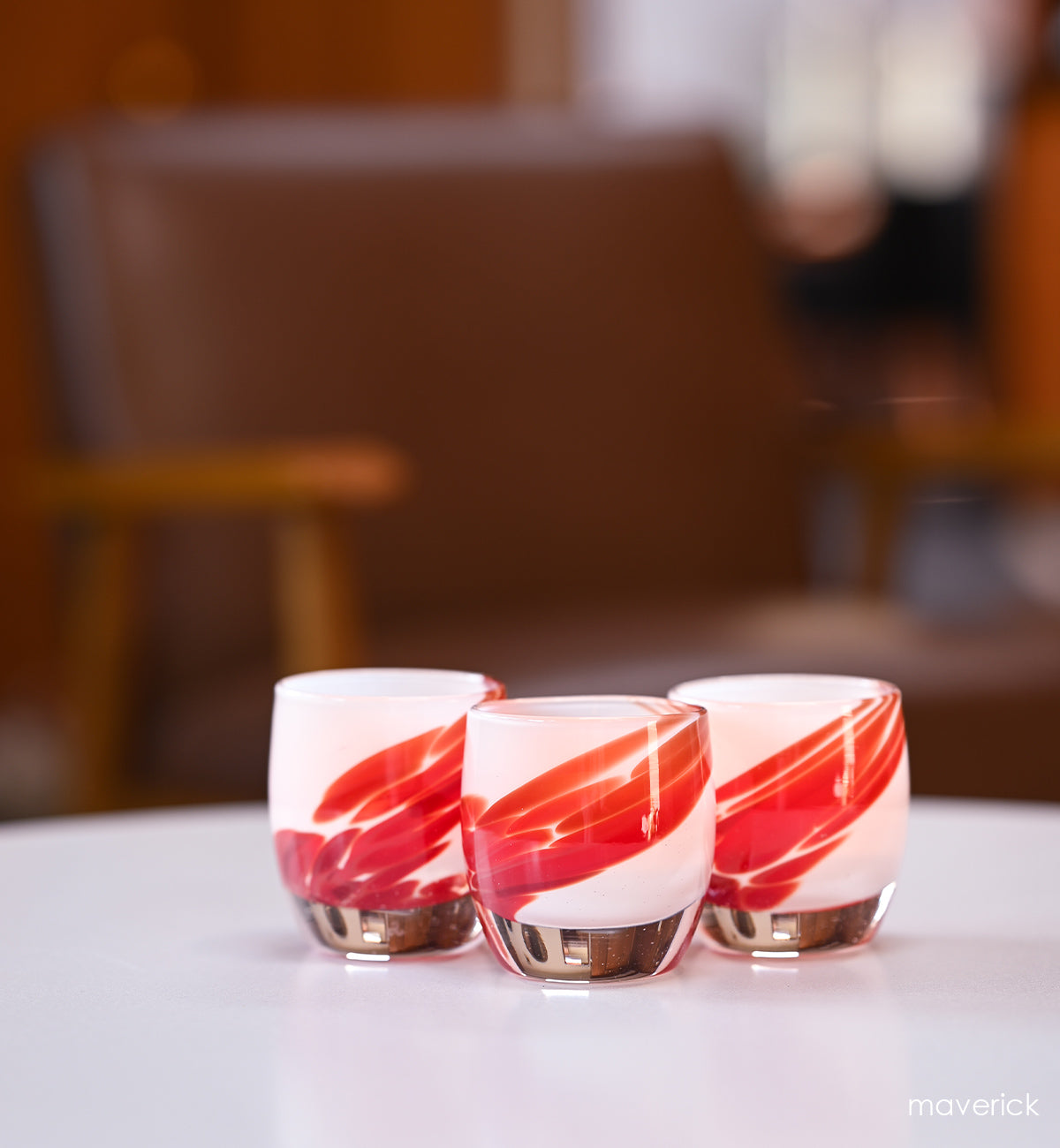 three maverick white with red wing, hand-blown glass votive candle holders on a white table with brown leather chair in background.