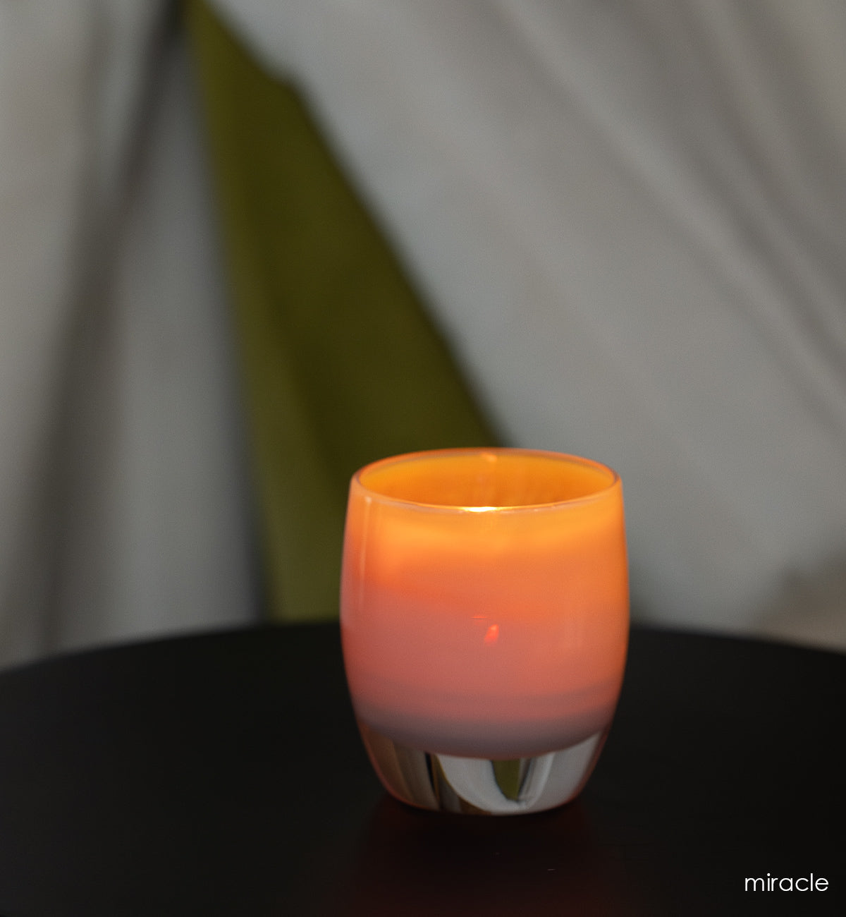 miracle golden pink hand-blown glass votive candle holder on dark table with white curtains in background.