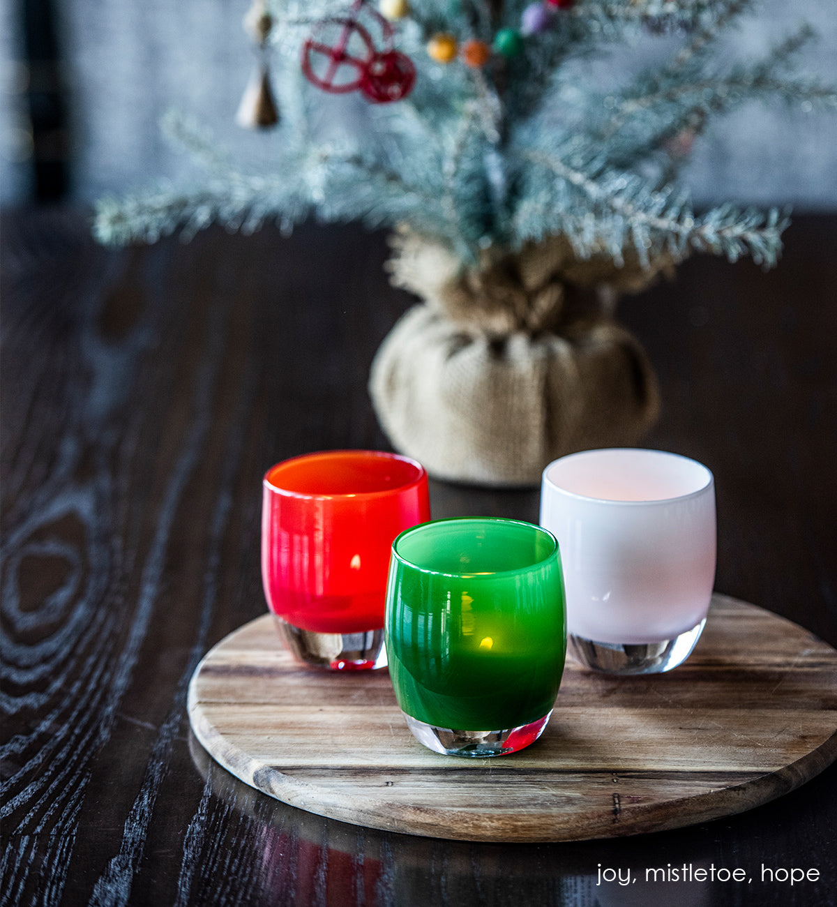 mistletoe green hand-blown glass votive candle holder. Paired with joy and hope on a wood circular table with decorative mini tree behind.