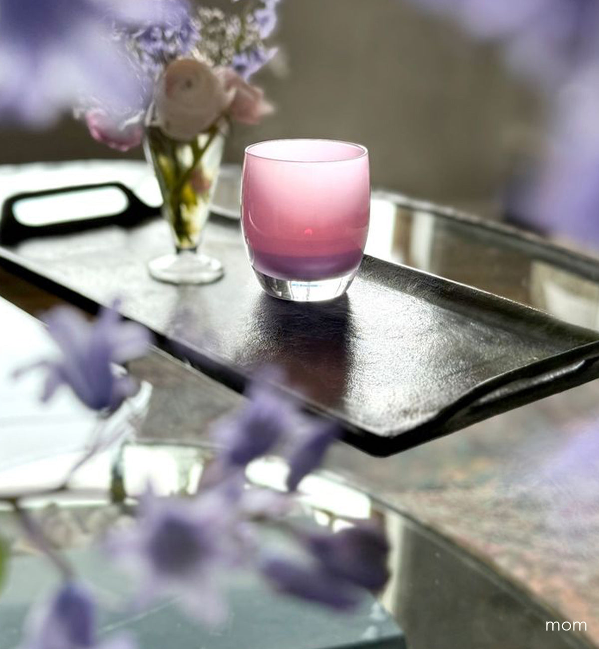 mom, soft purple hand-blown glass votive candle holder. on a dark metal tray with flowers sitting atop a glass coffee table with blurred surrounding purple flowers.