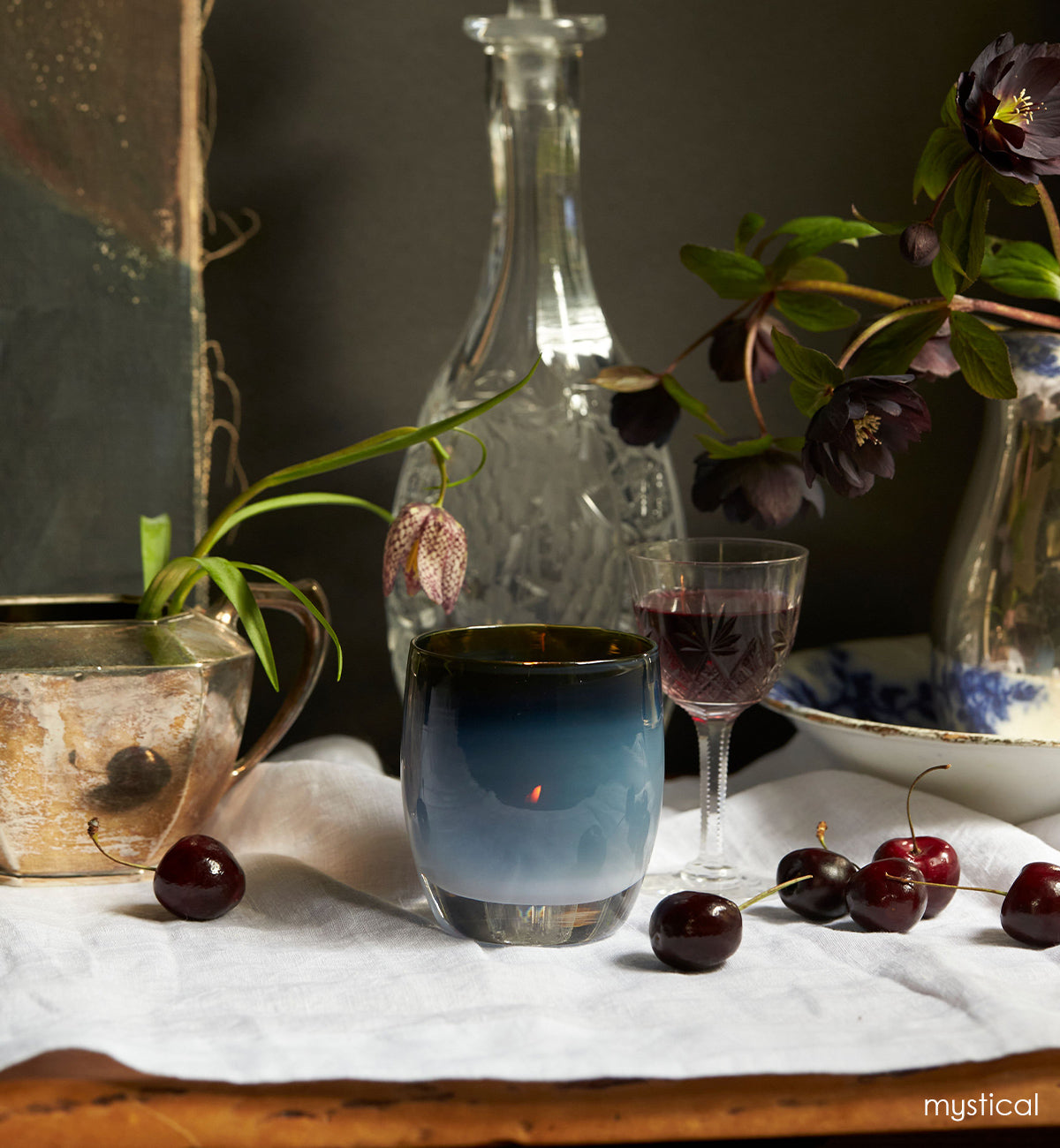 mystical, black opaline with metallic luster inside hand-blown glass votive candle holder on a table with cherries, wine and plants.