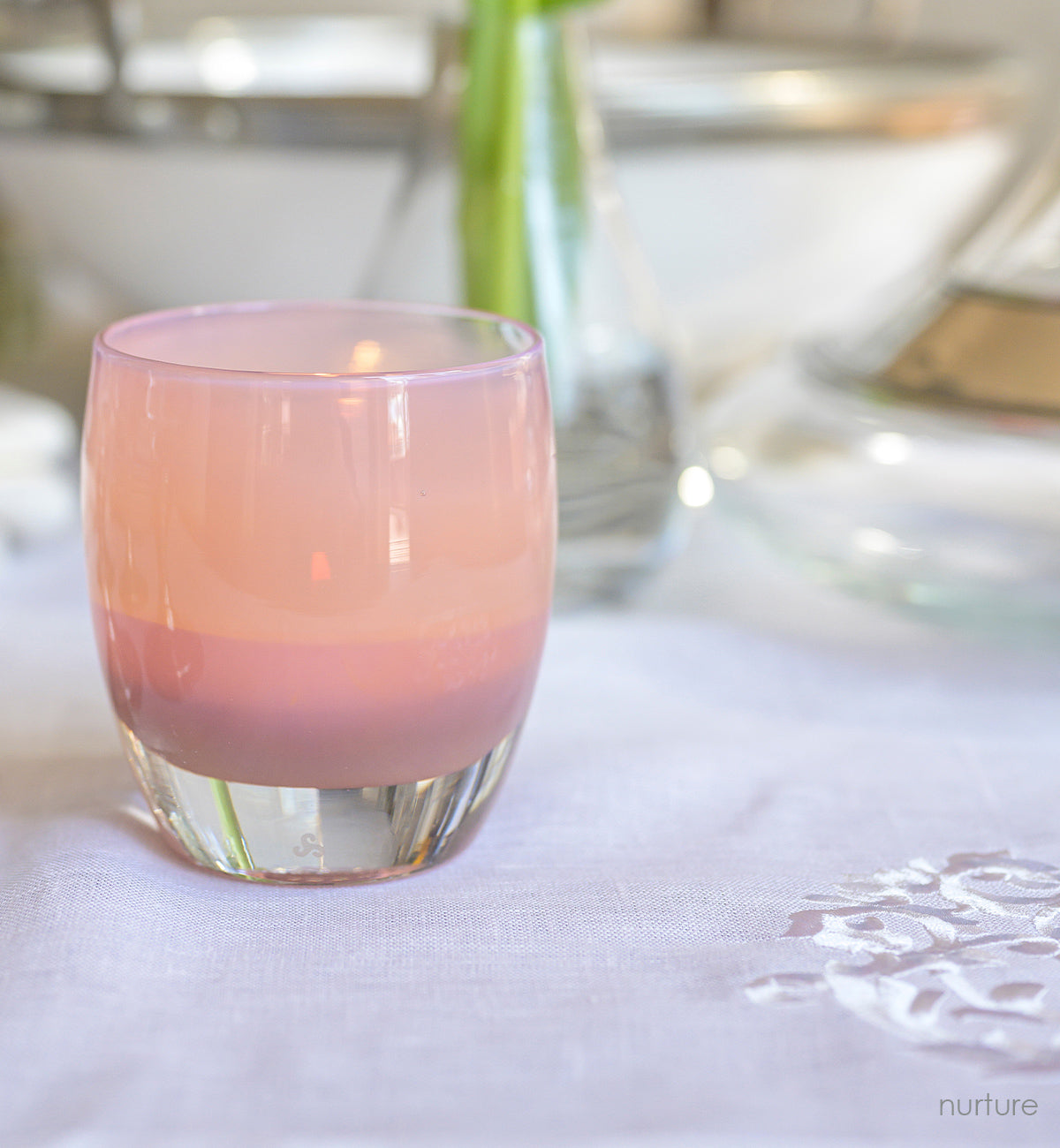 nurture, peachy glowing hand-blown glass votive candle holder, lit on a white tablecloth.