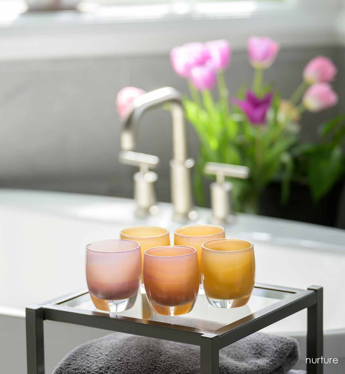 nurture, a peachy hand-blown glass votive candle holder, displayed next to a bathtub and fresh tulips.