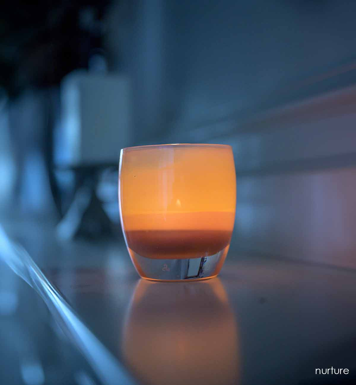 nurture, peachy glowing hand-blown glass votive candle holder, lit on a mantel.