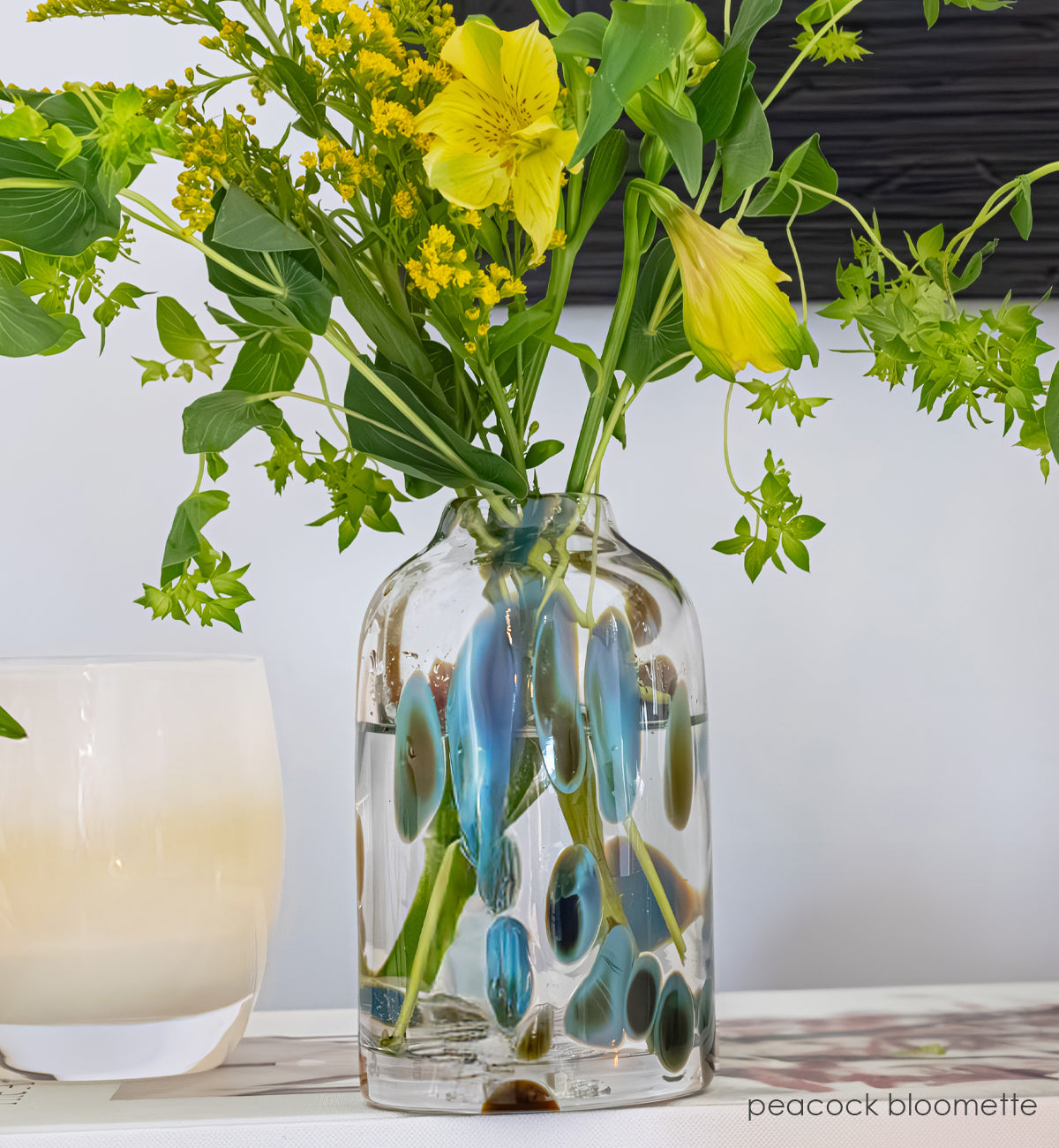 Clear glass vase with peacock pattern with yellow flowers and blue accents on a dark surface. angel glassybaby next to it.