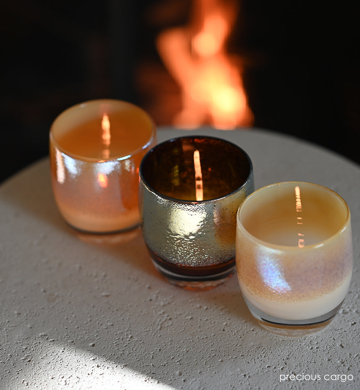 precious cargo is a beautiful set of 3 hand-blown glass votive candle holders; frankincense, gold, and myrrh sitting atop a concrete table surface with a fire blurred in the background.