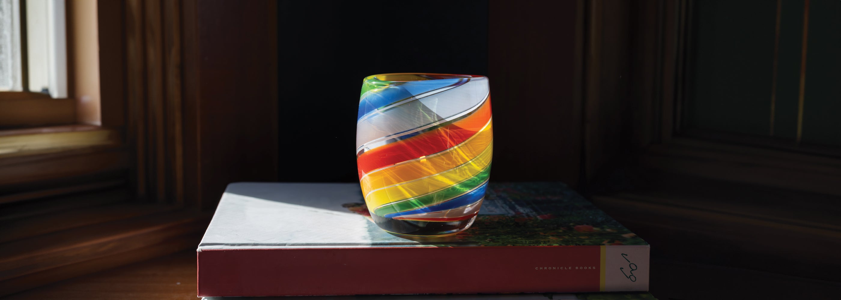 'pride 2025' glassybaby with rainbow swirls, hand-blown glass candle holder sitting on a book by a window.