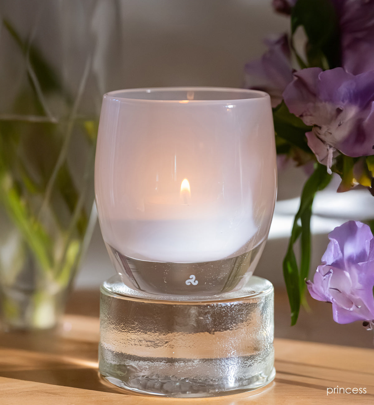 Soft pink candle in a glass holder with a blurred background of flowers.