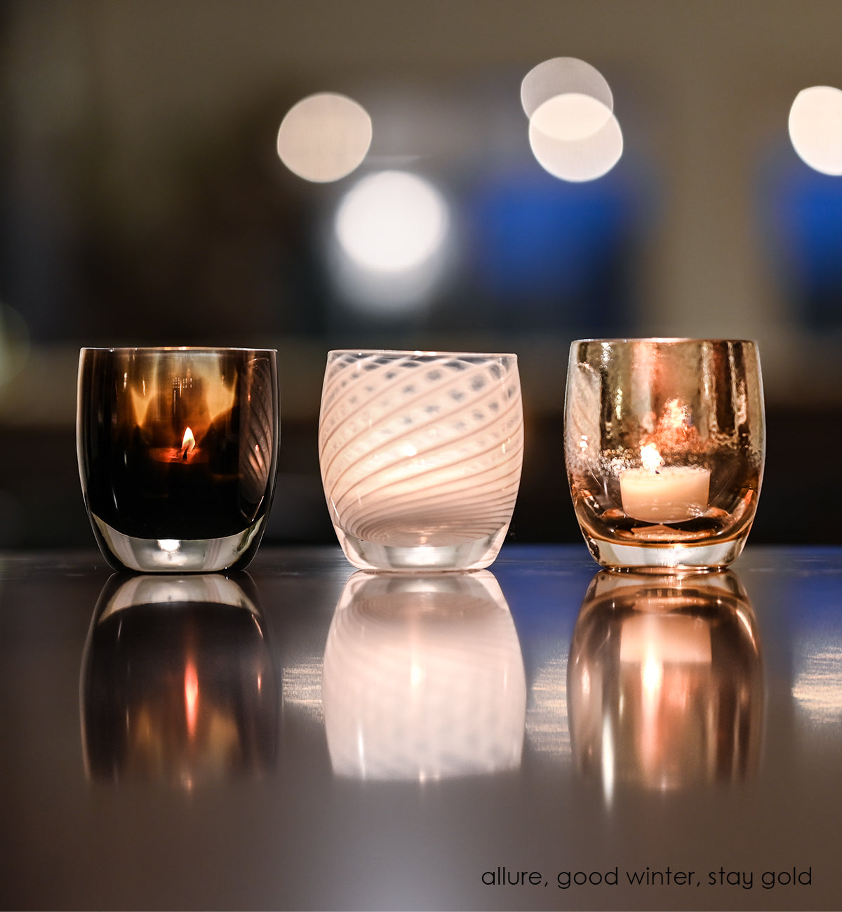allure, good winter, stay gold hand-blown glass votive candle holders lined up on a reflective table with lights blurred in background.