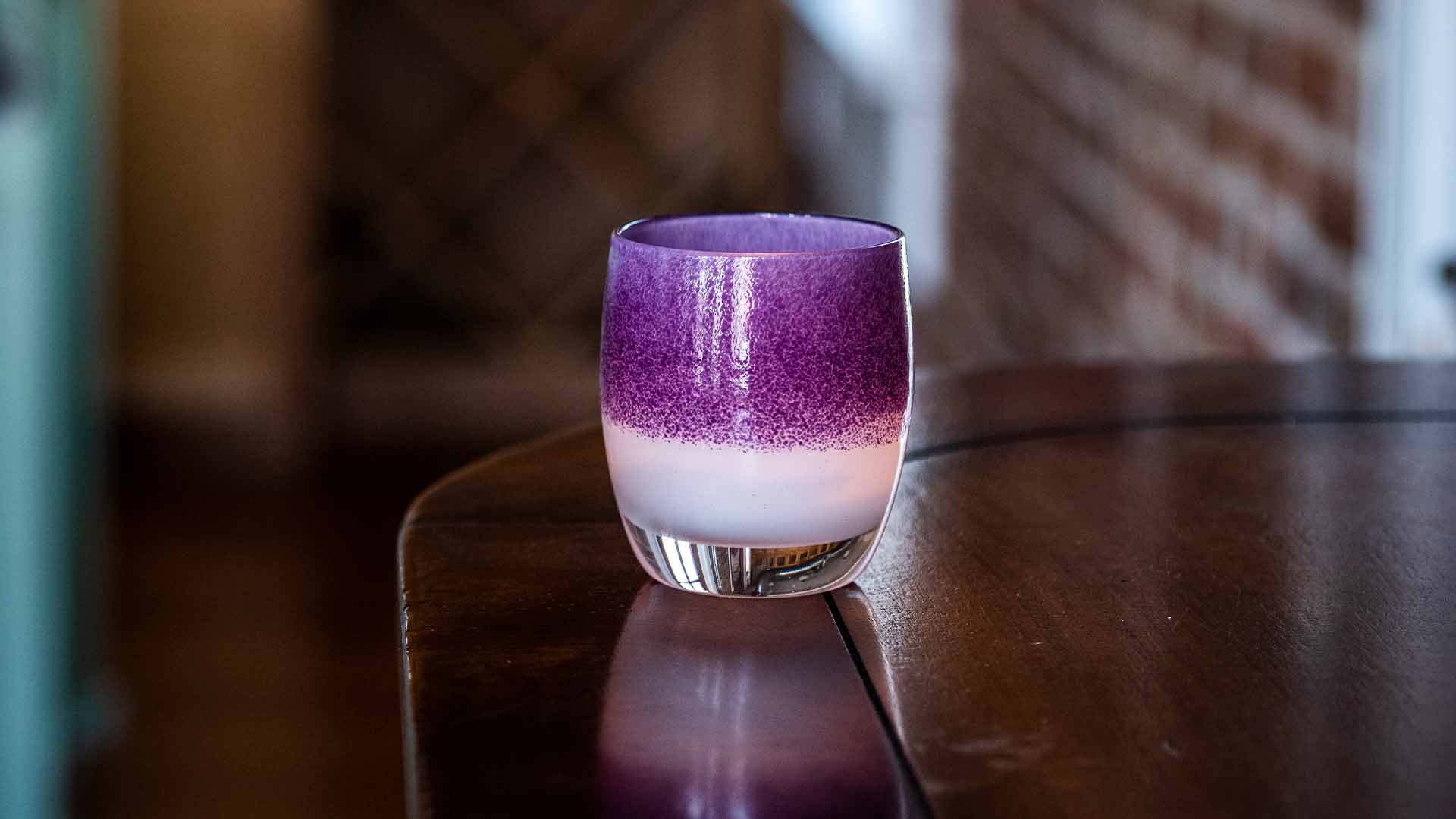 protector purple on white, hand-blown glass votive candle holder, sitting on a dark wood counter backed by exposed brick.
