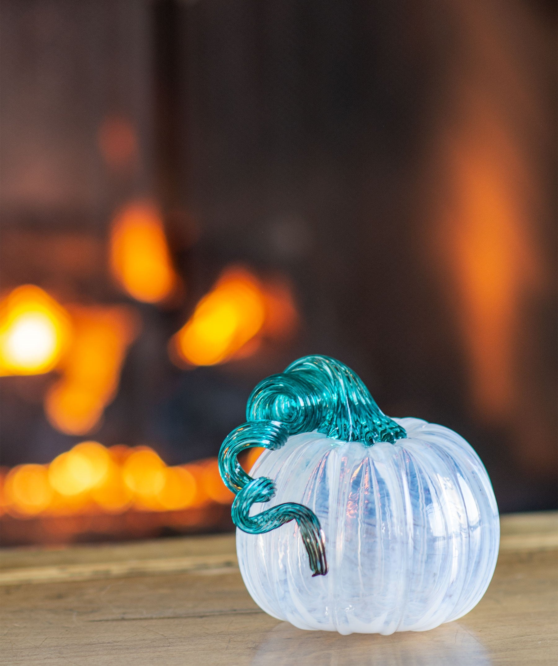 hand-blown glass pumpkin made in a glassybaby hot shop. with a white body and green stem sitting on a table in front of a lit fireplace.