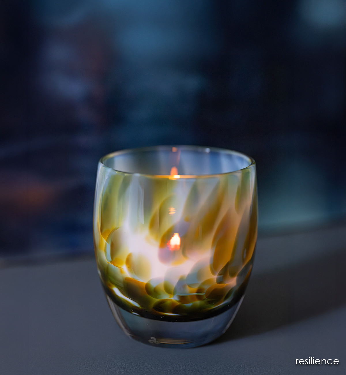 resilience | white hand-blown glass candle holder with brown, blue, and green petal deign with a lit candle against a dark background