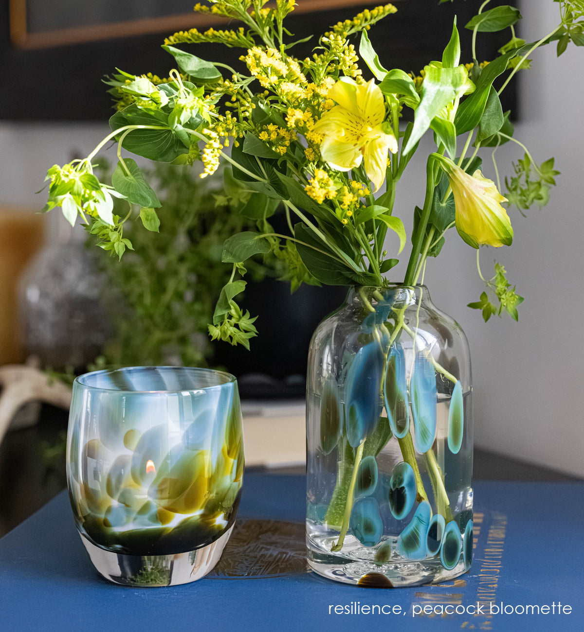 Clear glass vase with peacock pattern with yellow flowers and blue accents. with green and blue glassybaby next to it.