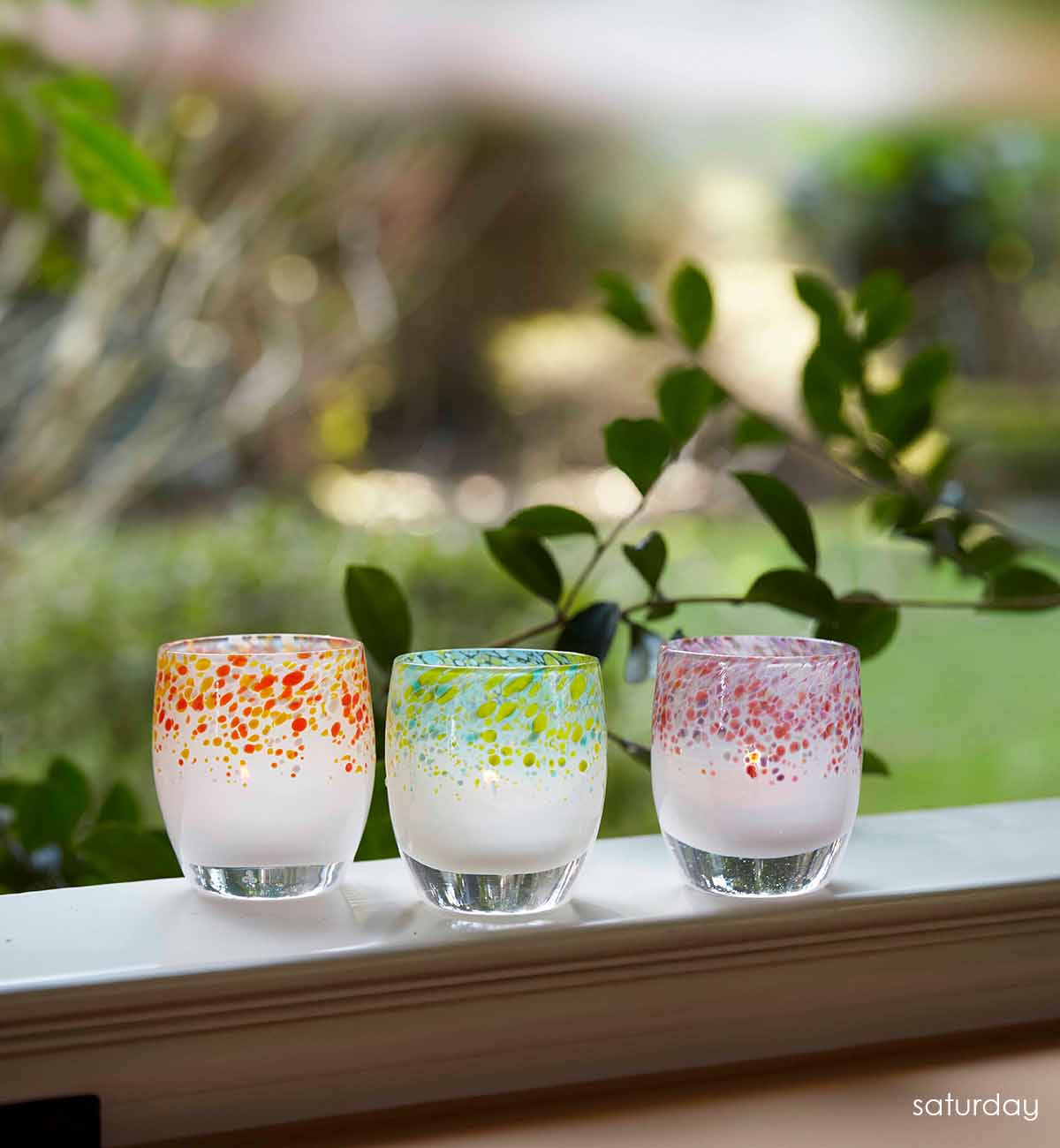 saturday warn yellow orange topped hand-blown glass votive candle holder, on a windowsill looking over a green yard.