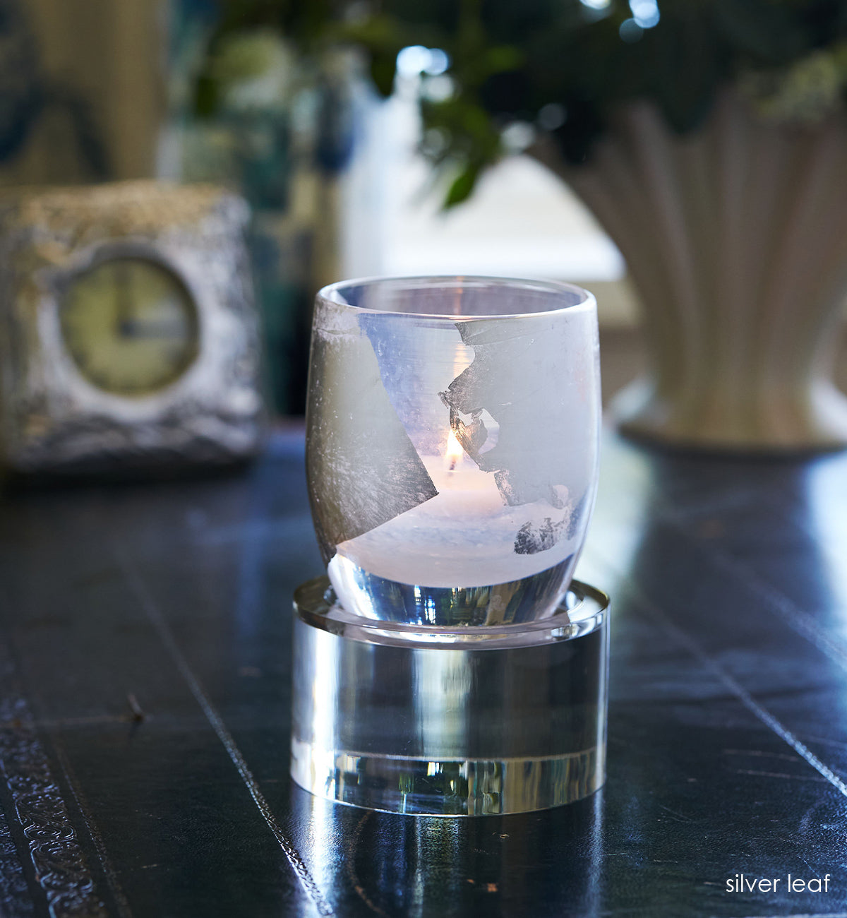 silver leaf, hand-blown glass votive candle holder with silver leaf, with a lit tealight glowing inside. sitting on a round baby stand on a navy blue desk with decorative clock and vase in background.