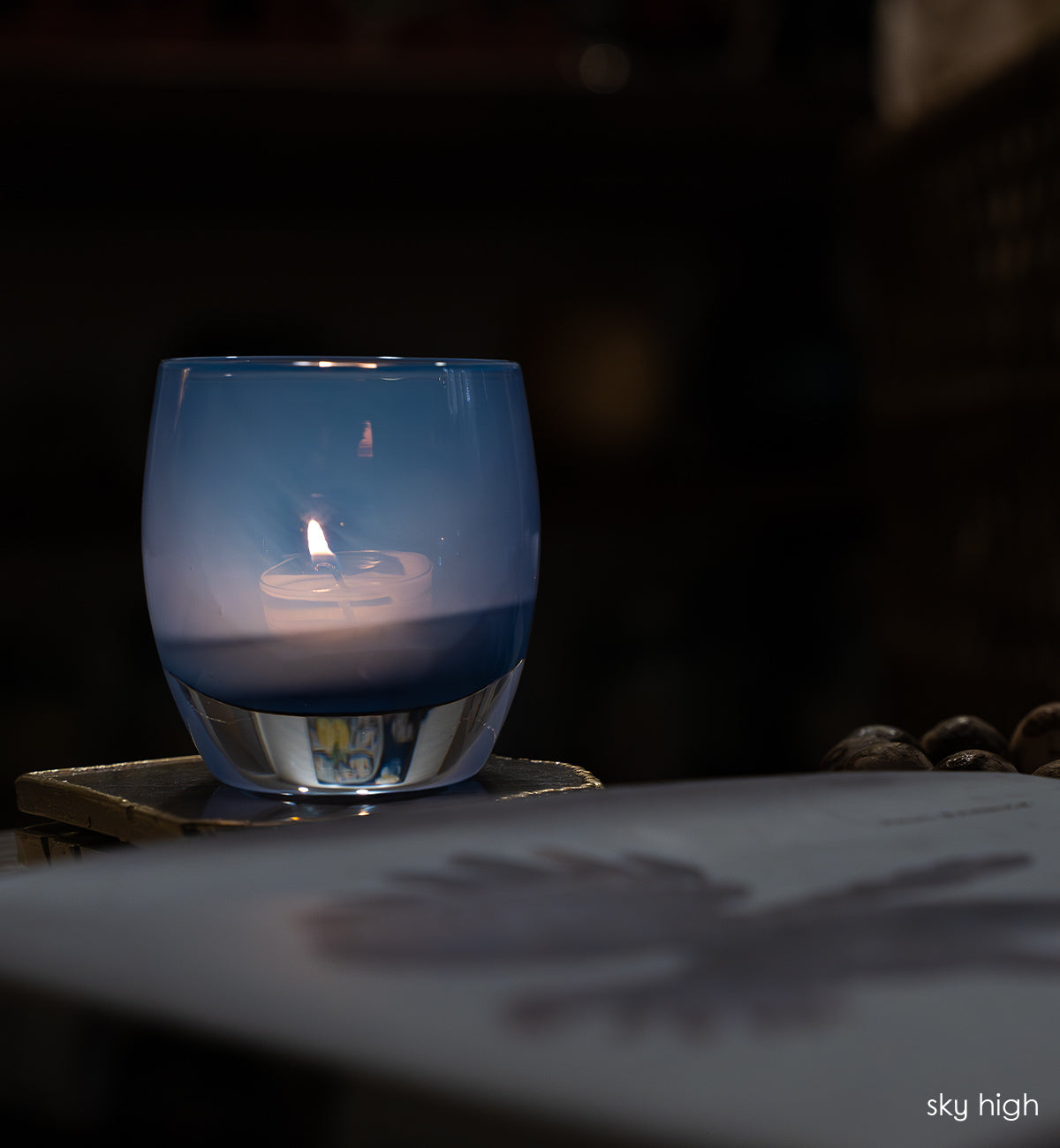 sky high blue hand-blown glass votive candle holder lit in a dark room on a crystal coaster.