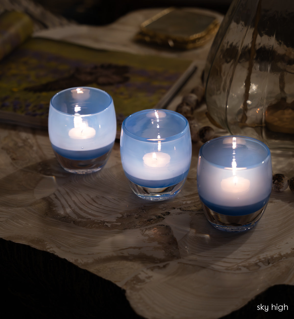 three sky high blue hand-blown glass votive candle holders on a patterned table cloth alongside a glass vase, crystal coasters and coffee table book