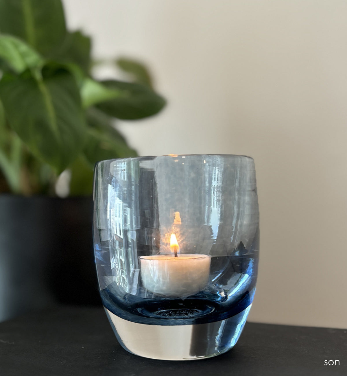 lit 'son' blue hand-blown glass candle holder on black book. Green plant in black pot behind it.