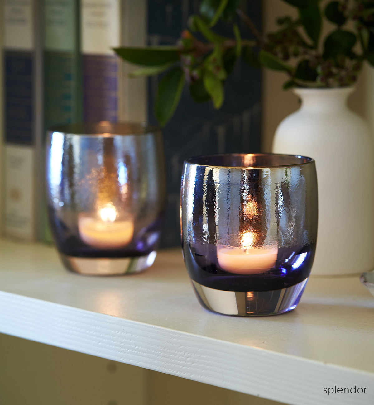 two splendor, purple with silver shimmer hand-blown glass votive candle holders on a white bookshelf with books and vase behind.