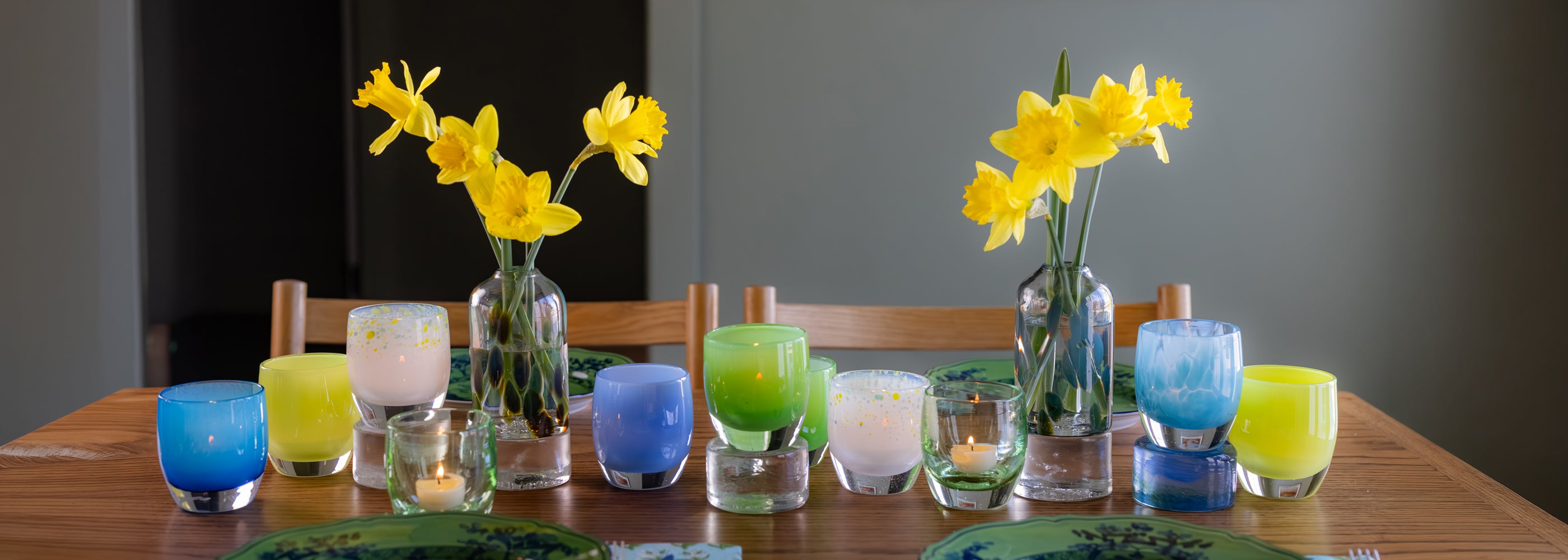 Table setting with colorful spring glass candle holders and bloomette vases with yellow flowers on a wooden table.
