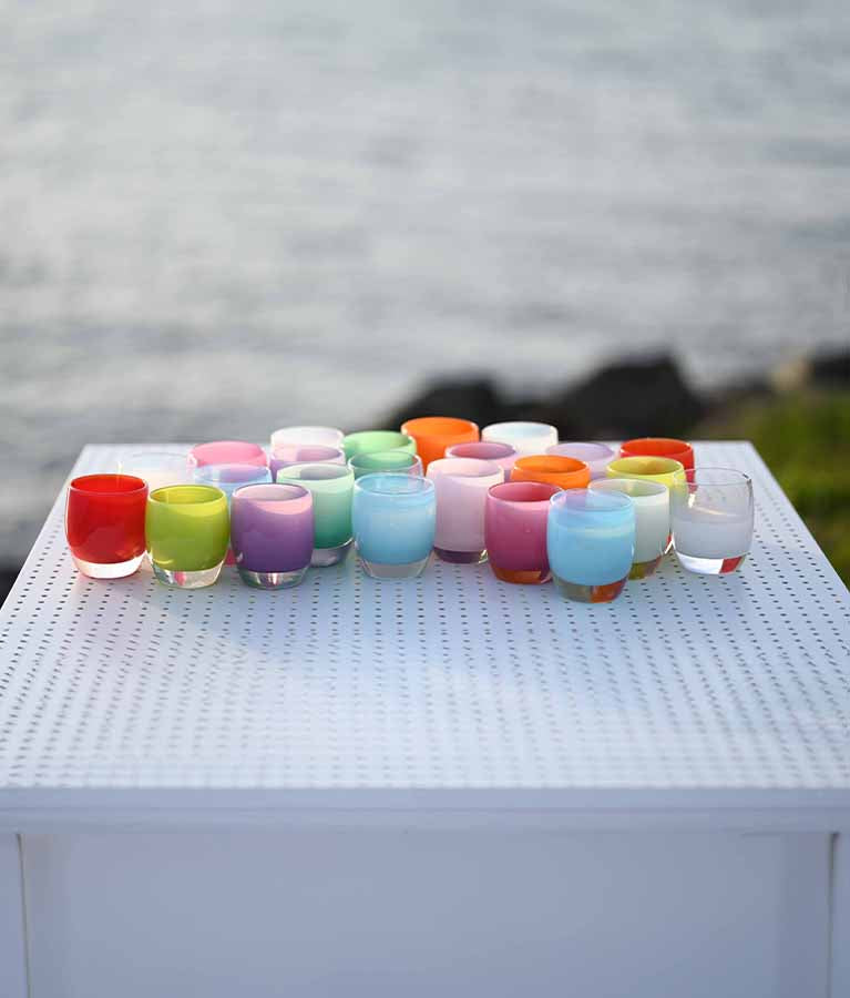 spring collection, an array of colored hand-blown glass votive candle holders on a white table overlooking the water.