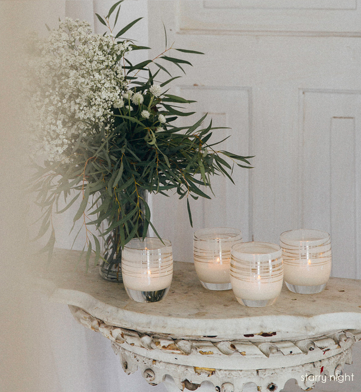 four starry night, white with silver shimmer wrap hand-blown glass votive candle holders sitting on a tall rustic table with a vase of white flowers and green leaves in front of a white door.
