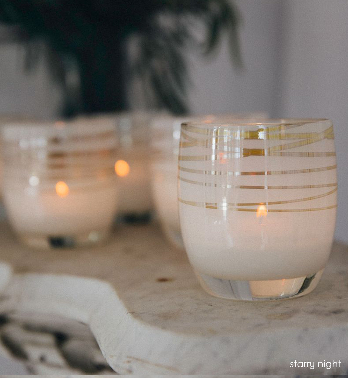 grouping of starry night, white with silver shimmer wrap hand-blown glass votive candle holders lit on a rustic surface with green plant blurred in background.