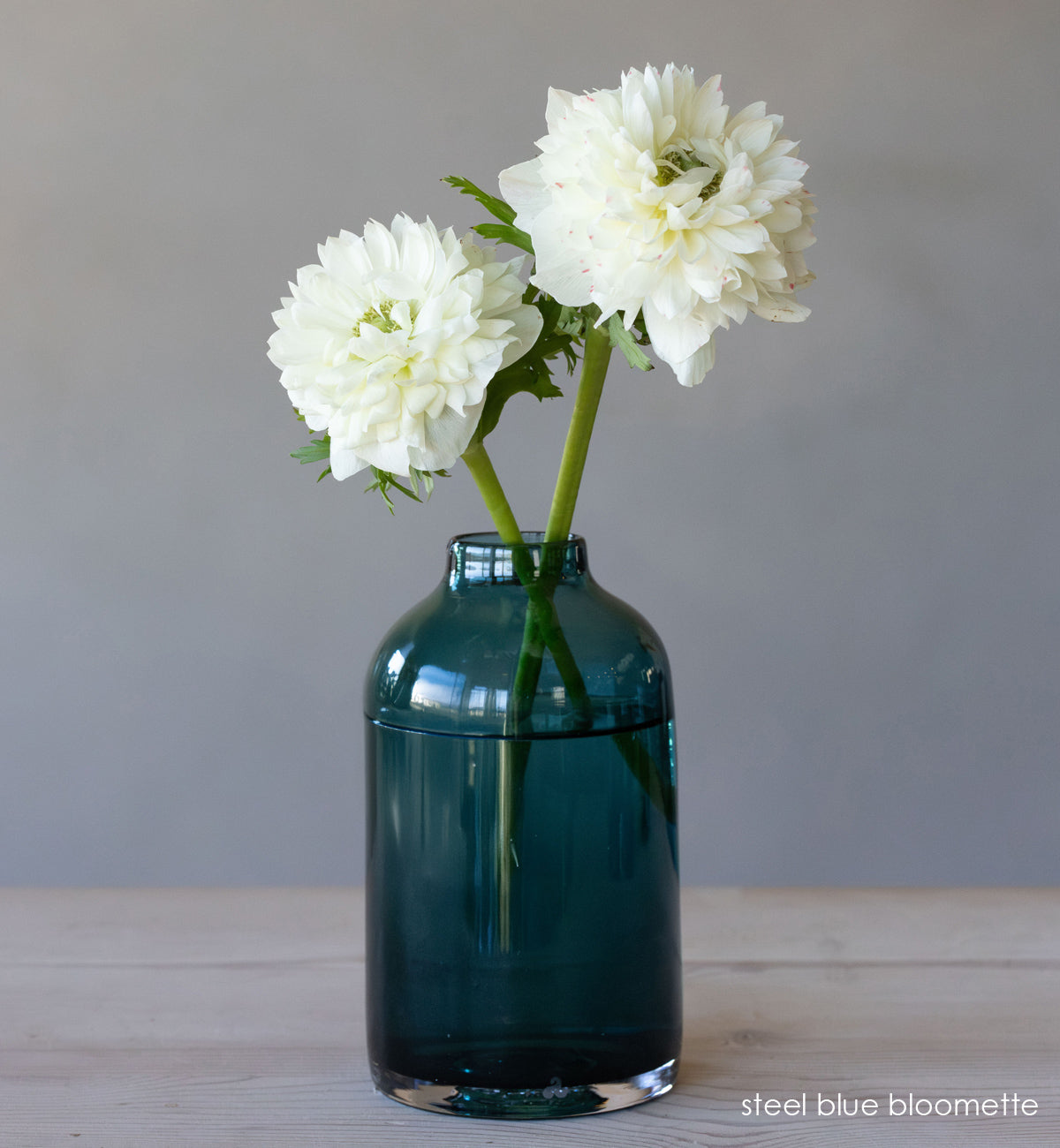 transparent blue hand-blown glass flower bud vase with white flowers on wood table and gray background