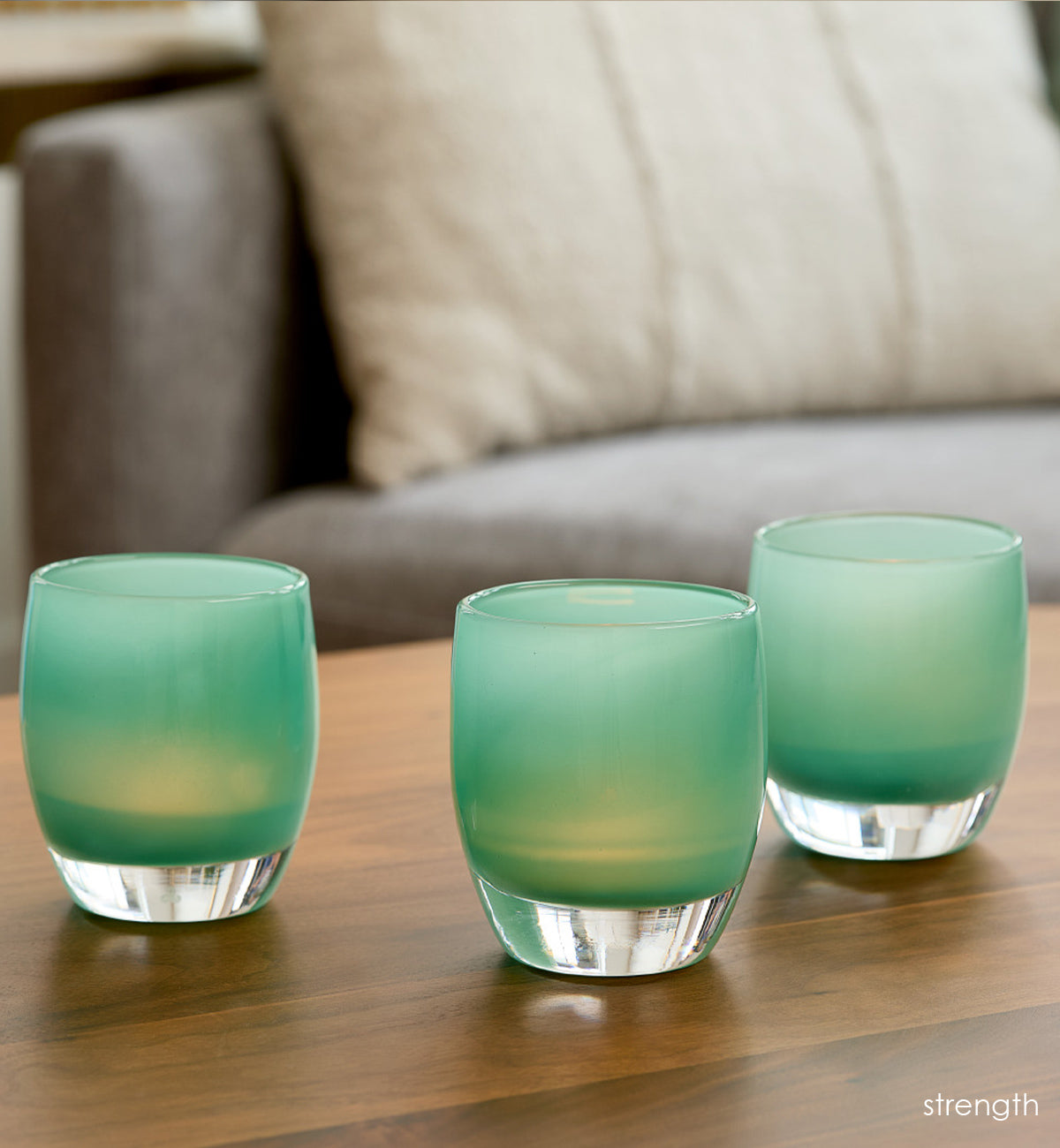 three strength teal green hand-blown glass votive candle holders on a wood coffee table with gray couch and cream pillow blurred in background.