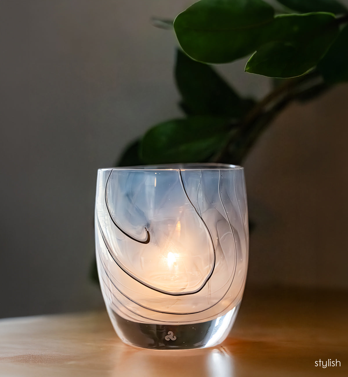 white with black line design glass candle holder with a lit candle on a wooden surface, with a plant in the background.