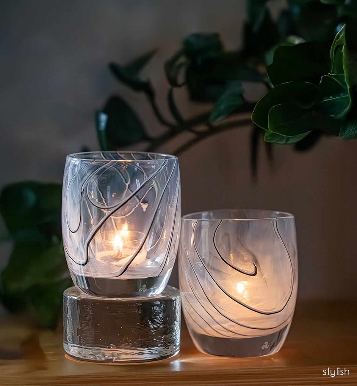 Two glass candle holders with a subtle black line design, one on top of a pedestal next to the other, on a wooden surface with a plant in the background.