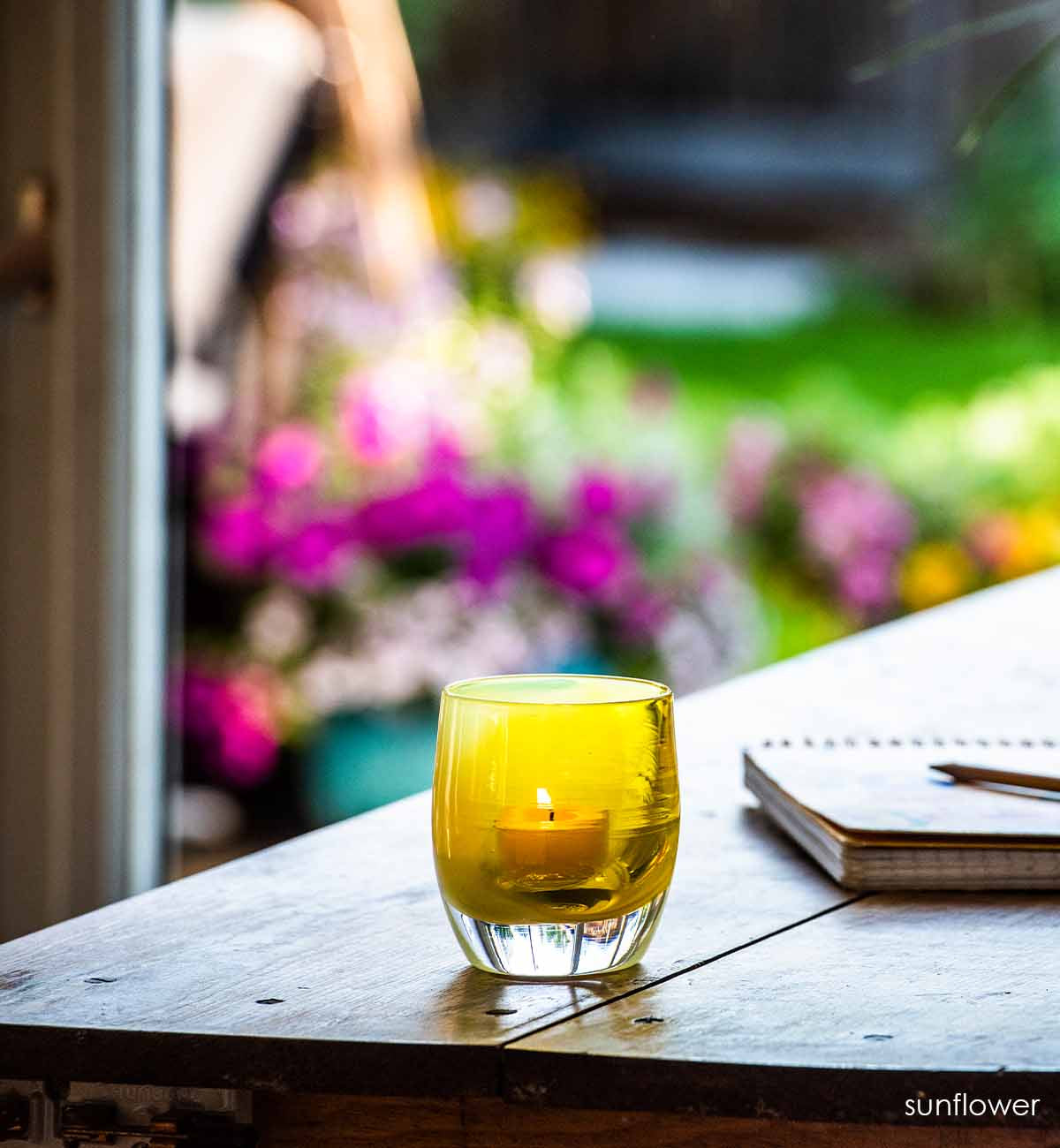 sunflower on a wooden table looking over a floral garden. two toned yellow hand-blown glass votive candle holder.