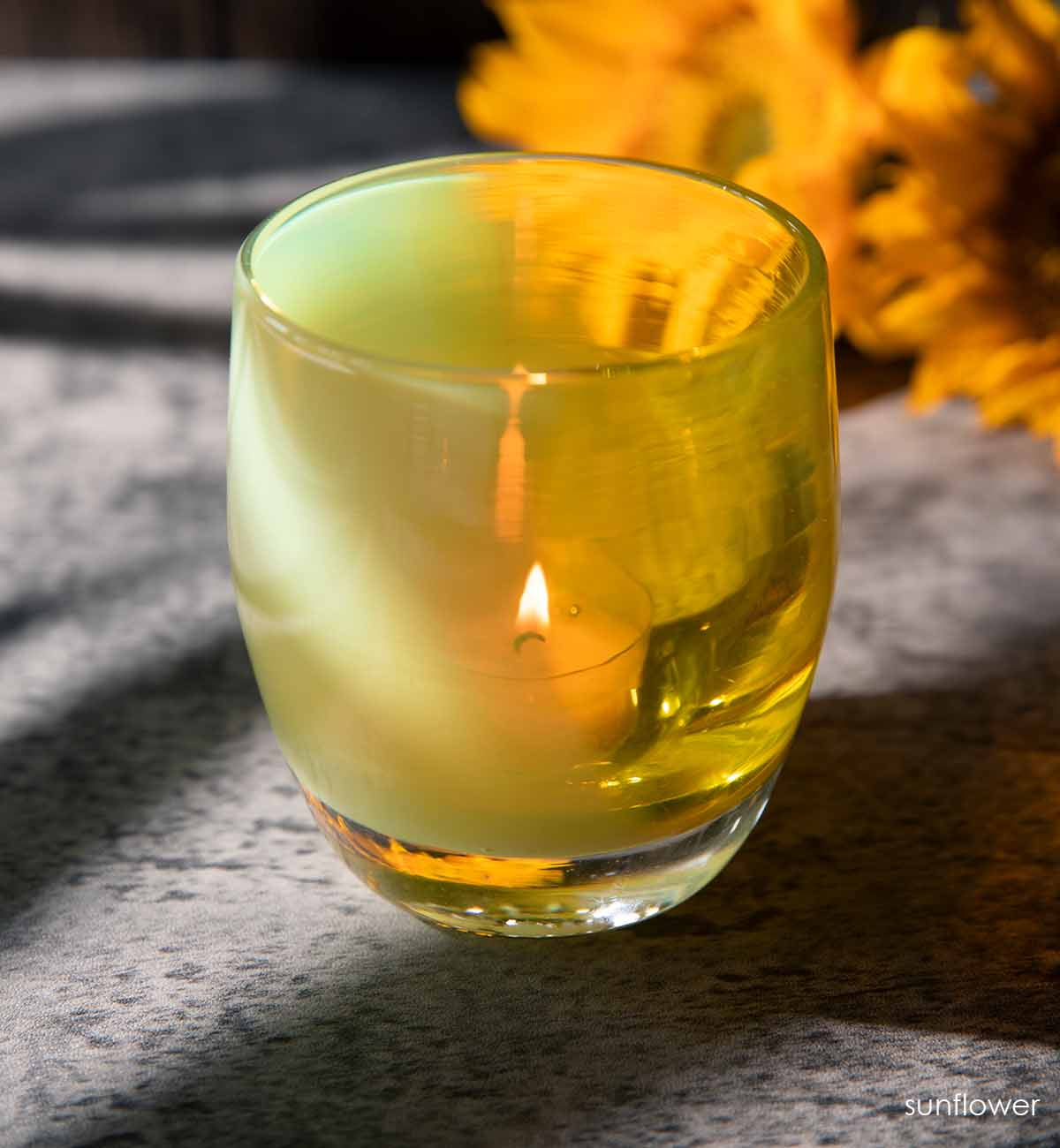 sunflower dual textured yellow glass votive candle holder, casting shadows in the sun next to a bundle of sunflowers.