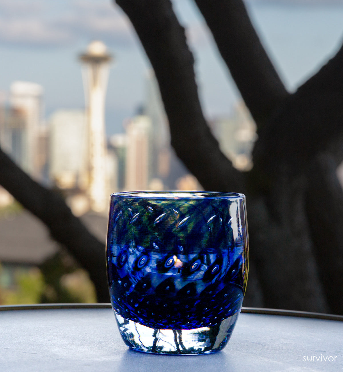 survivor sapphire blue bubble, hand-blown glass votive candle holder sitting on circular table in front of a blurred tree and Seattle space needle and skyline background.